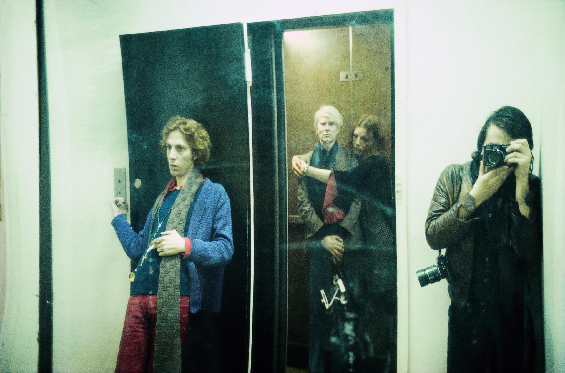 Fotographie von vier Personen durch einen Spiegel. Ein Mann lehnt außen an der Tür eines Aufzugs. Im Aufzug steht Andy Warhol, der von einer Frau umarmt wird. Beide blicken in die Kamera. Rechts vor dem Aufzug steht ein Mann dessen Gesicht von einer Kamera verdeckt ist. 